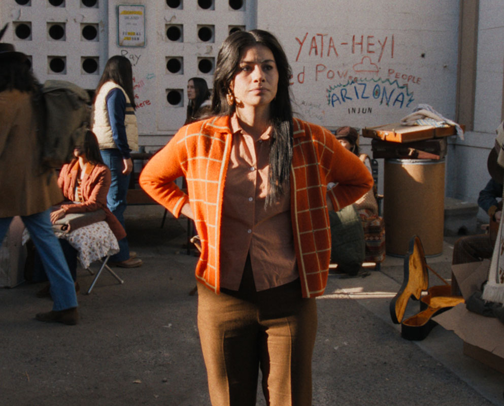 A Native woman in an orange cardigan stands with her hands on her hips in a crowded courtyard, beneath a wall painted with “Red Power,” in a scene from Episode 8 of People of the West.