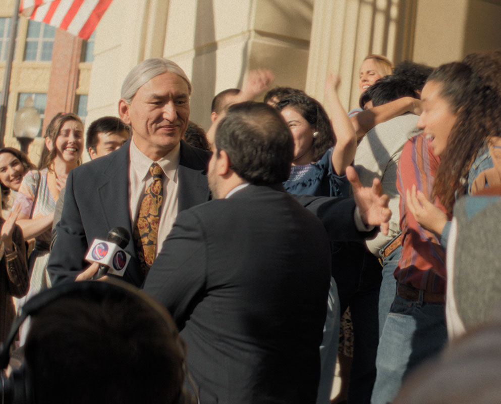 A Native man in a suit smiles at a reporter’s microphone as a crowd celebrates outside a courthouse, in a scene from People of the West.