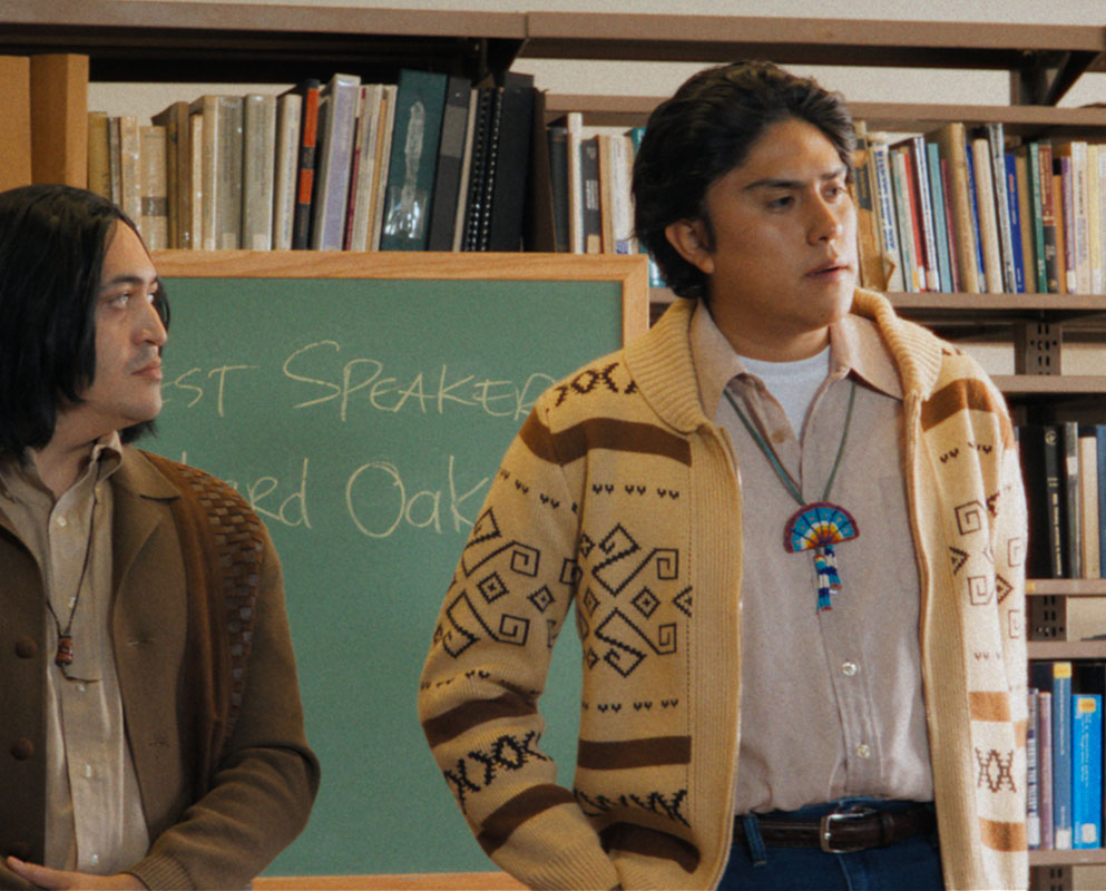 Two young men in 1960s clothing stand in a library beside a chalkboard announcing guest speaker Richard Oakes, in a scene from Episode 8 of People of the West.