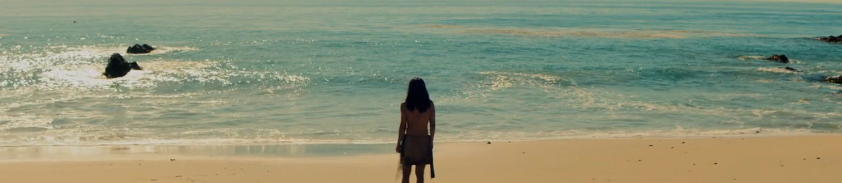 A Native person with long dark hair stands on a sandy beach with her back to the camera, looking out at the ocean, in a scene from People of the West.