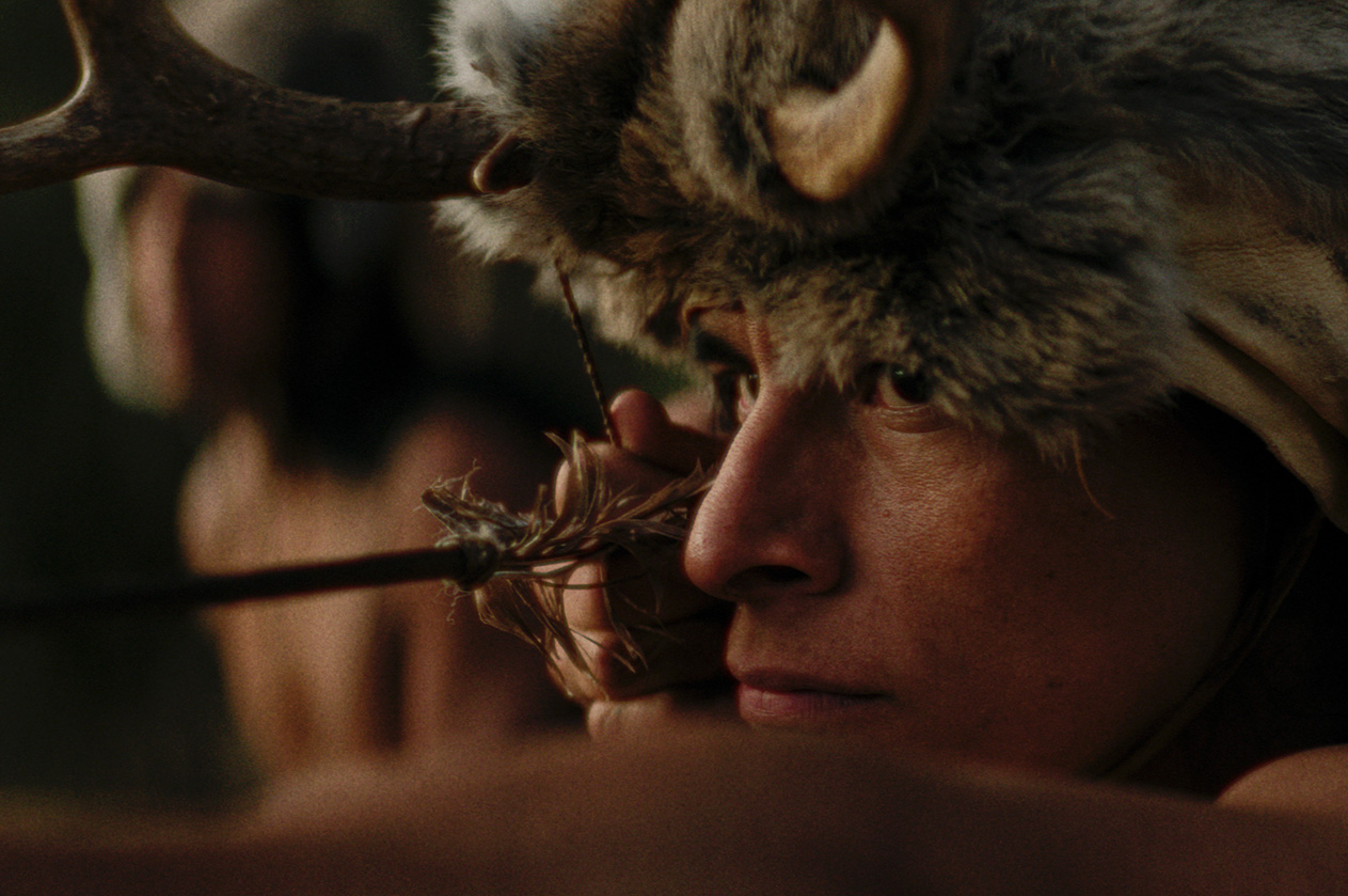 A Native hunter wearing a deer hide and antler headdress draws a feathered arrow on his bow, in a scene from People of the West.