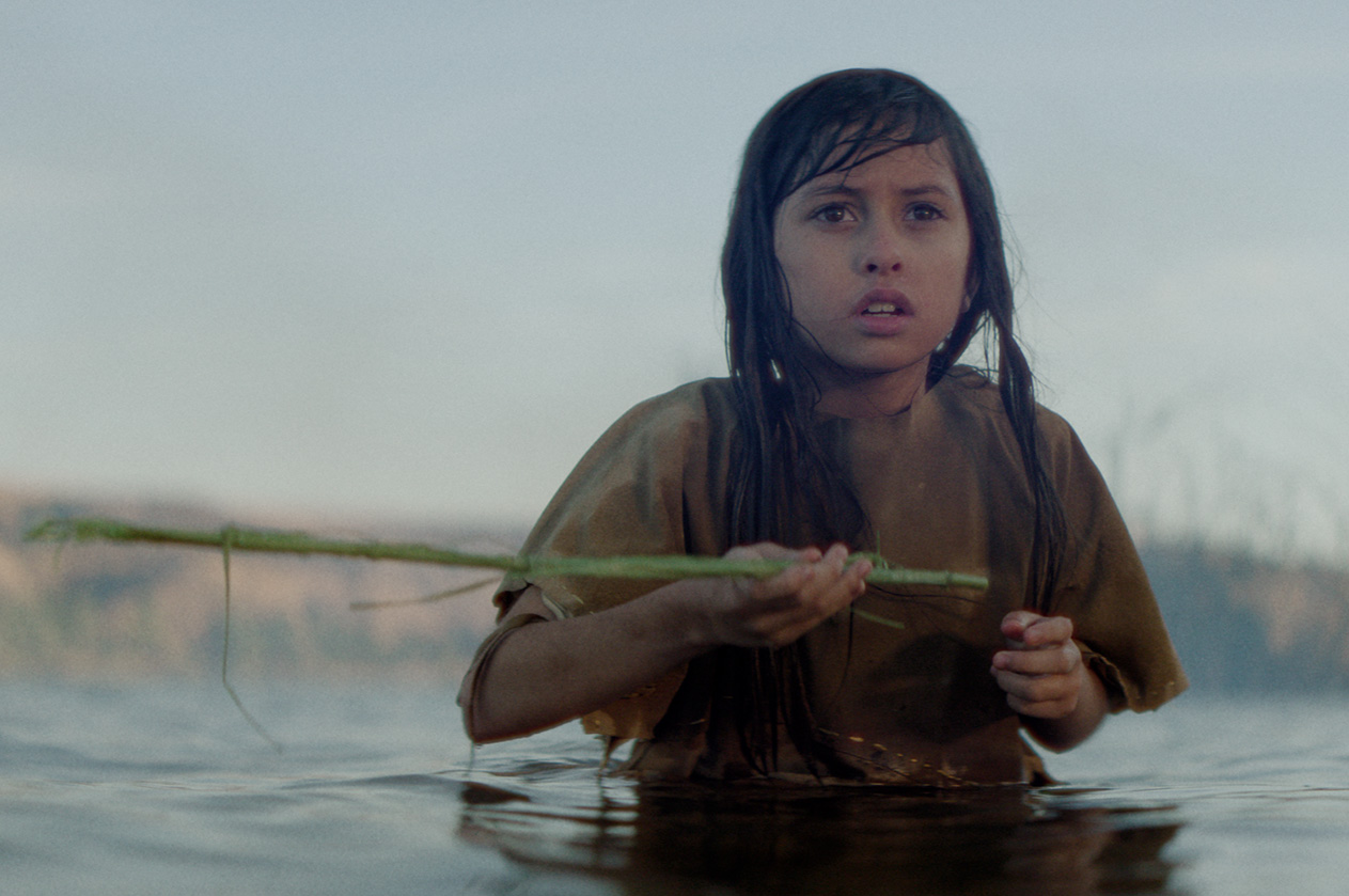 A Native girl stands waist deep in a river holding a green reed, looking ahead with alarm, in a scene from People of the West.