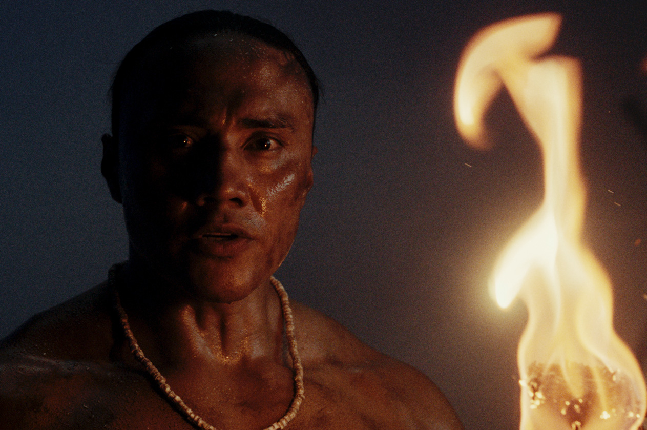 A Native man wearing a shell bead necklace stands in the dark, his face lit by a nearby flame, in a scene from People of the West.
