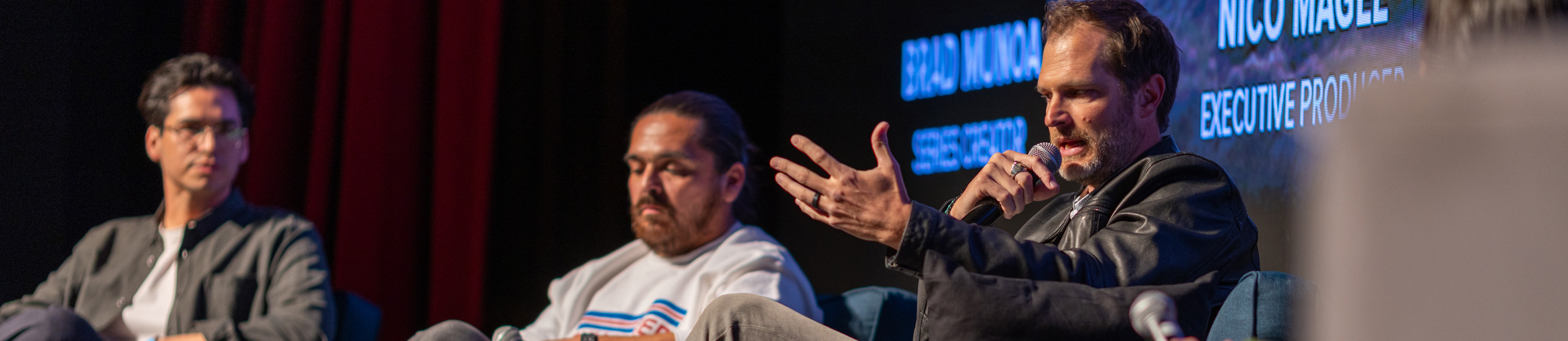 People of the West series creator Brad Munoa speaks into a microphone during a panel discussion, seated beside two fellow panelists on stage.