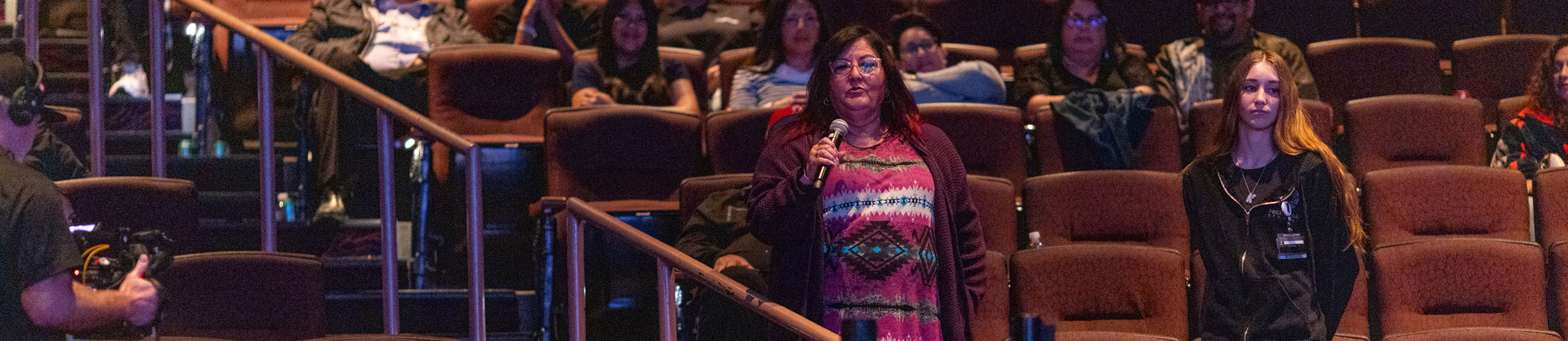 A woman stands in a theater aisle and speaks into a microphone during an audience Q&A at a People of the West screening.