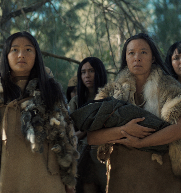 Native women and girls in fur and hide clothing stand together in a forest, one holding a bundle wrapped in a blanket, in a scene from People of the West.
