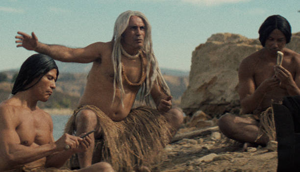 Three Native men in grass skirts sit on a rocky shoreline as an elder with long white hair gestures toward the water, in a scene from People of the West.