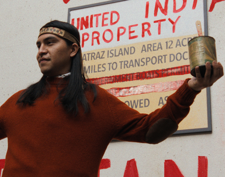 A young Native man in a headband holds up a paint can beside the Alcatraz Island sign repainted with “Indian,” in a scene from Episode 8 of People of the West.