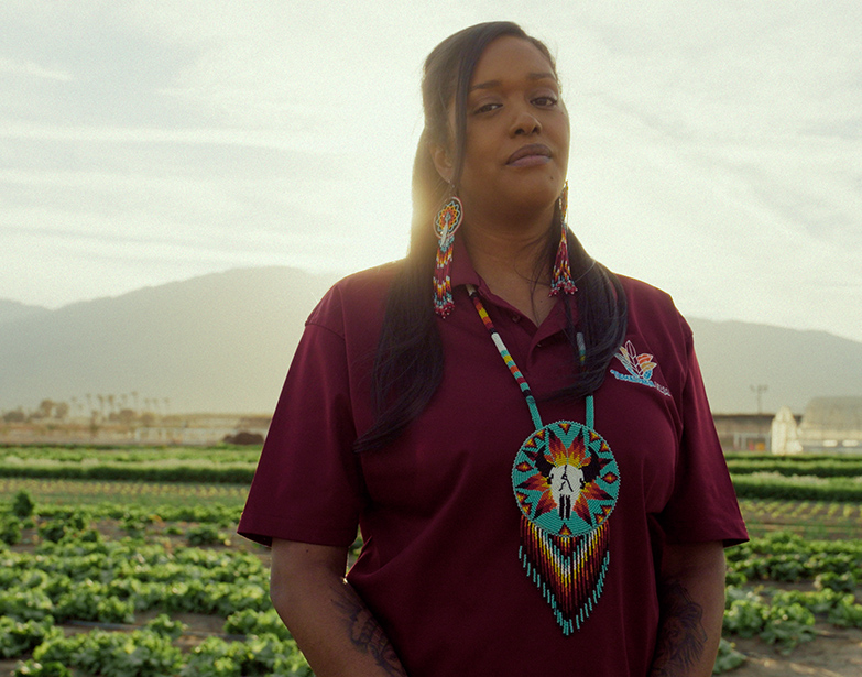 A Native woman wearing a large beaded medallion and beaded earrings stands in a farm field at sunset, in People of the West.