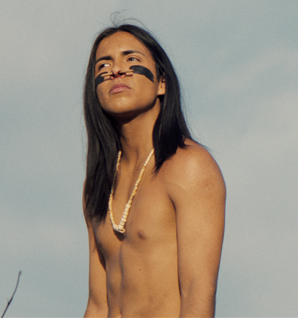 A young Native man with black face paint and a shell bead necklace looks out against an open sky, in a scene from People of the West.