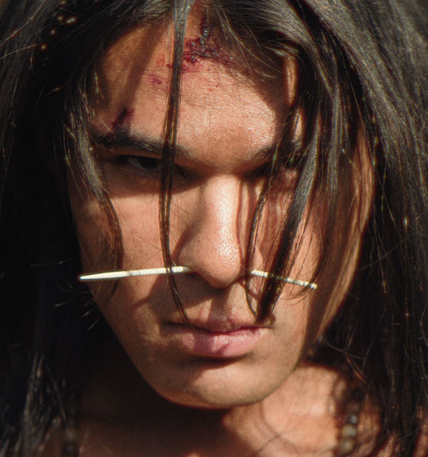 A young Native man with long hair, a nose piercing, and a cut on his forehead stares intently, in a scene from People of the West.