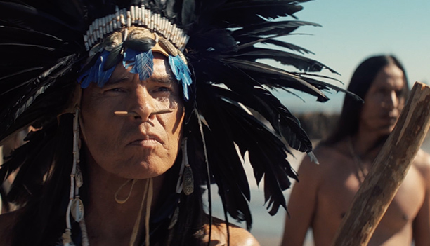 A Native leader in a tall black feather headdress with a nose piercing stares ahead on a shoreline as a man holding a wooden staff stands behind him, in a scene from People of the West.