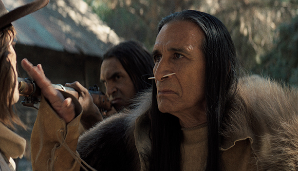 A Native elder in a fur cloak, with long dark hair and a nose piercing, stands firm as a man in a hat points a rifle beside him, in a scene from People of the West.