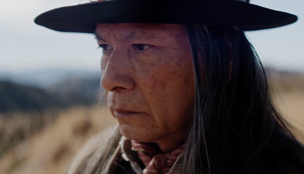 A Native elder in a black wide-brimmed hat, with long graying hair, gazes out across dry hills, in a scene from People of the West.