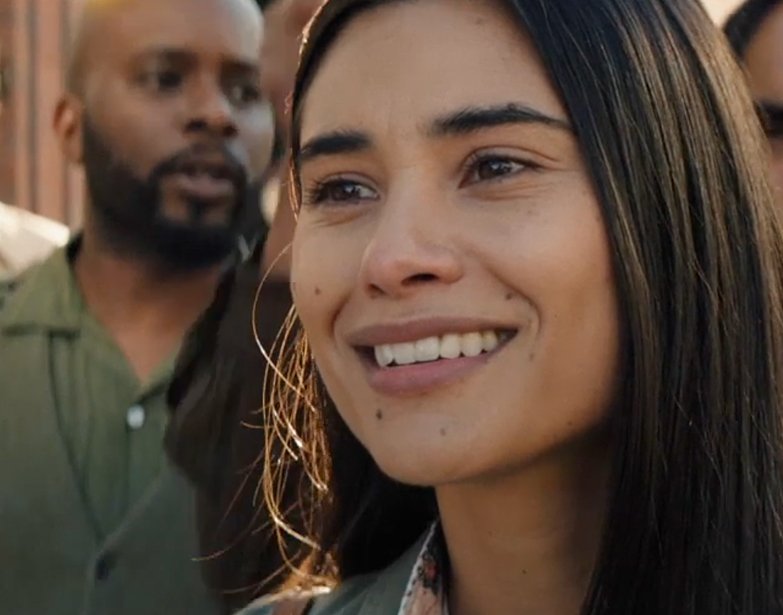 A Native woman with long dark hair and hoop earrings smiles in a crowd, in a scene from People of the West.
