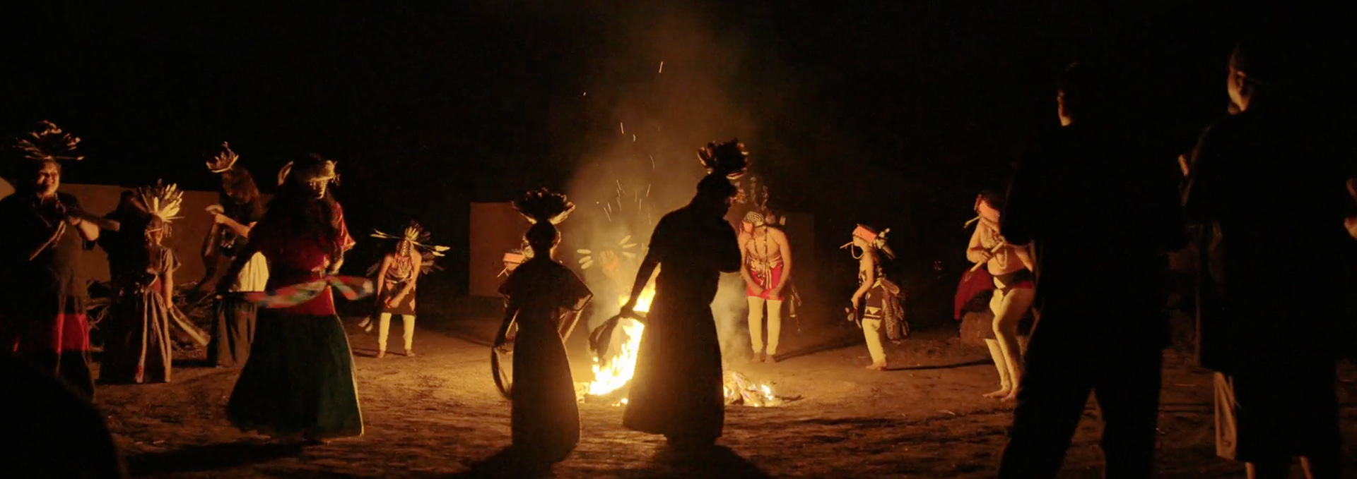 Native dancers in feather headdresses and ceremonial regalia dance in a circle around a bonfire at night, in a scene from People of the West.