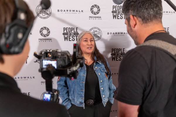 A woman with long gray hair, wearing a denim jacket, is interviewed by a camera crew in front of a People of the West backdrop with Pechanga Studios and Boardwalk Pictures logos.